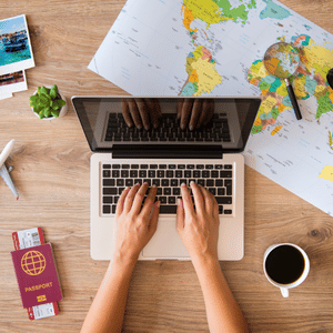 Hands typing on laptop beside world map, passport, coffee, and travel photos on a wooden desk