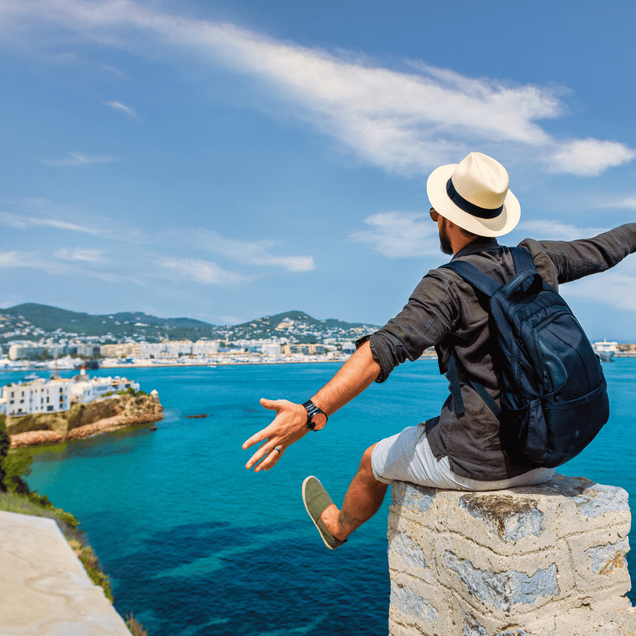 Traveler with backpack and hat sitting on a stone ledge overlooking a turquoise bay and coastal town