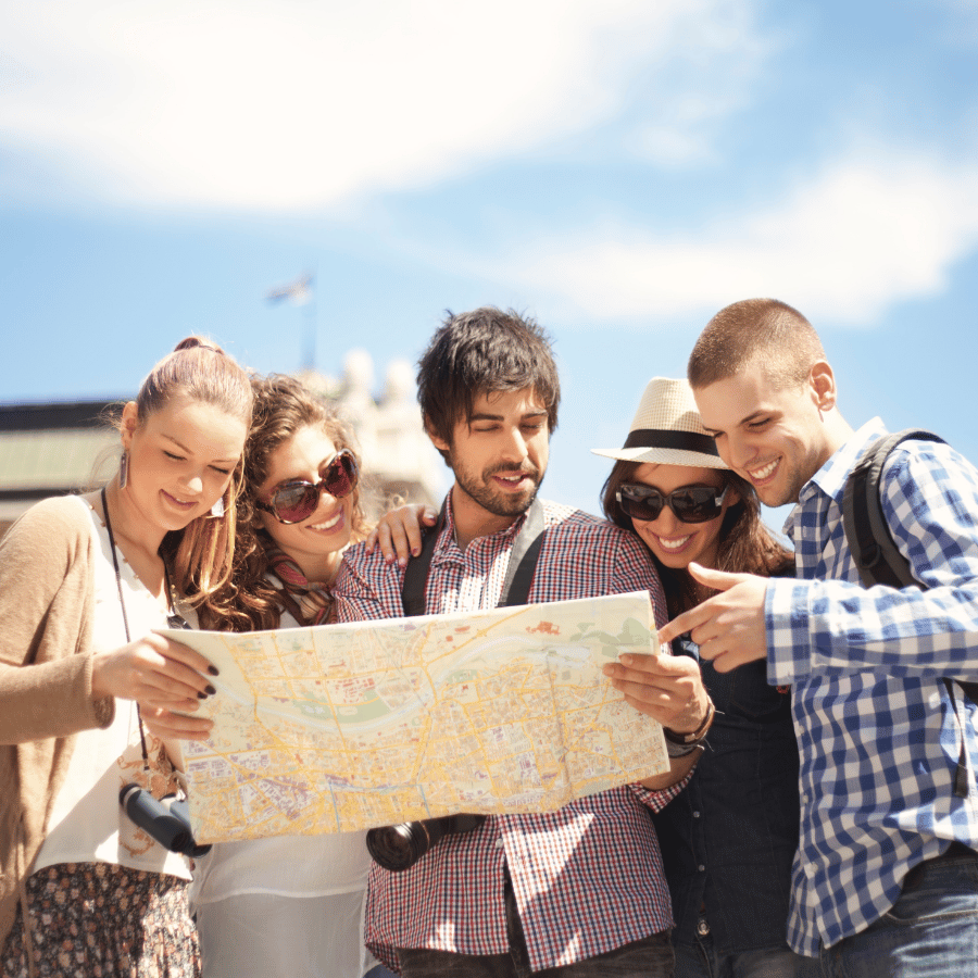 Five friends outdoors studying a map under a blue sky, smiling and pointing directions