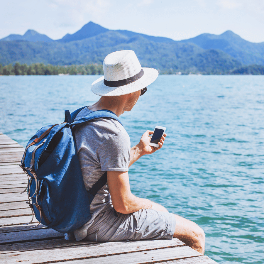 Person sitting on a dock by a blue lake, looking at a phone, with mountains in the background