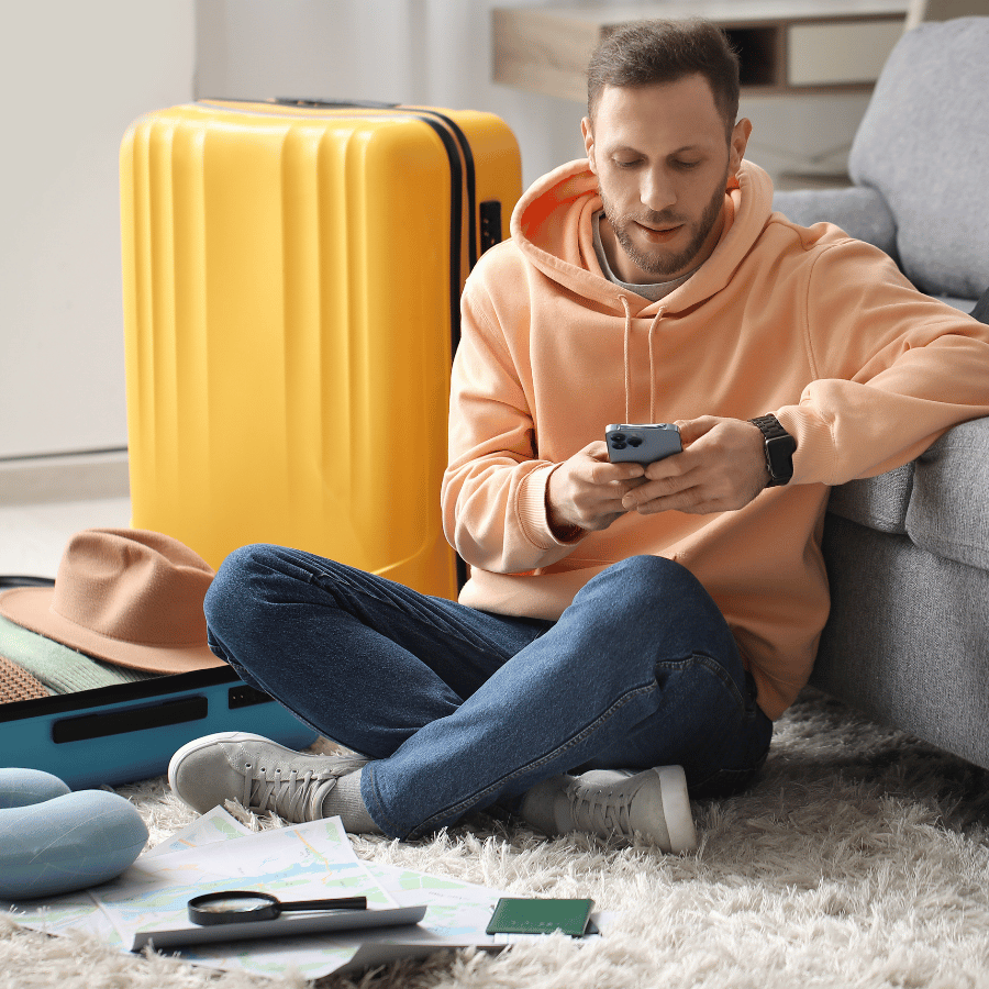 Man sitting on the floor beside an open yellow suitcase, looking at his phone in a living room.