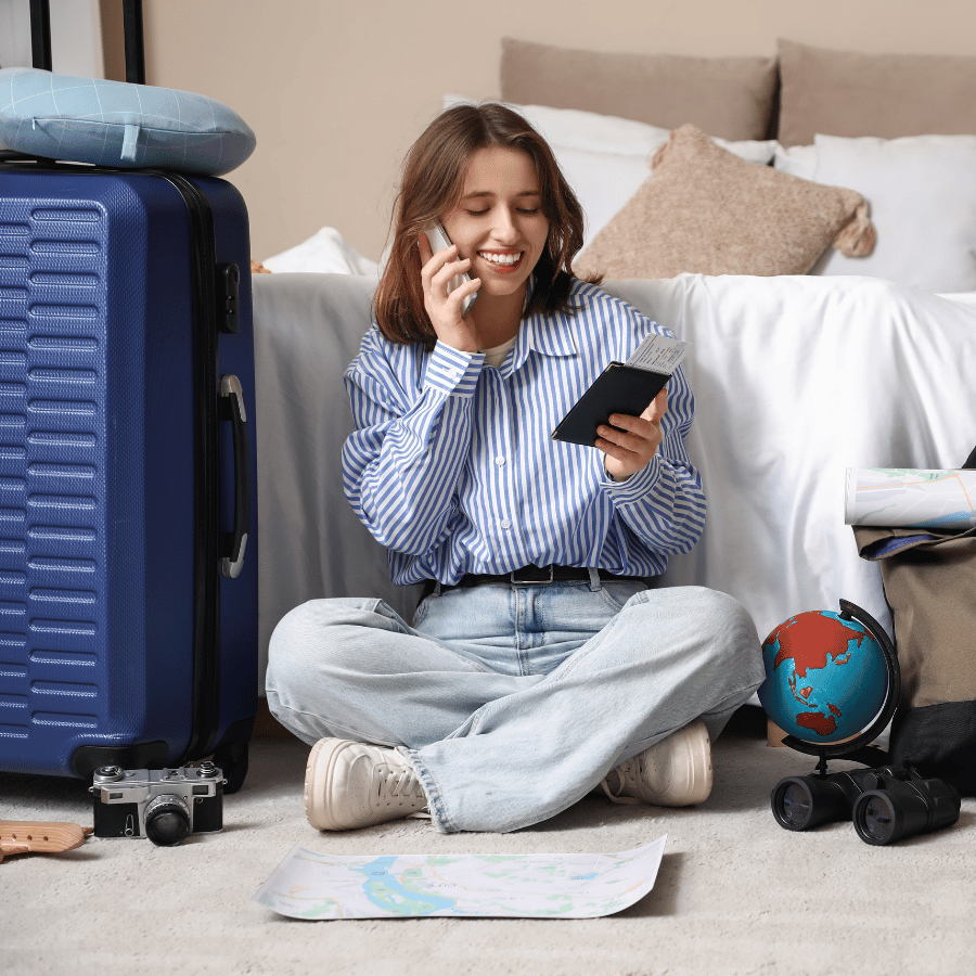 Person sitting on the floor in a bedroom, smiling at a phone beside open luggage and travel items.