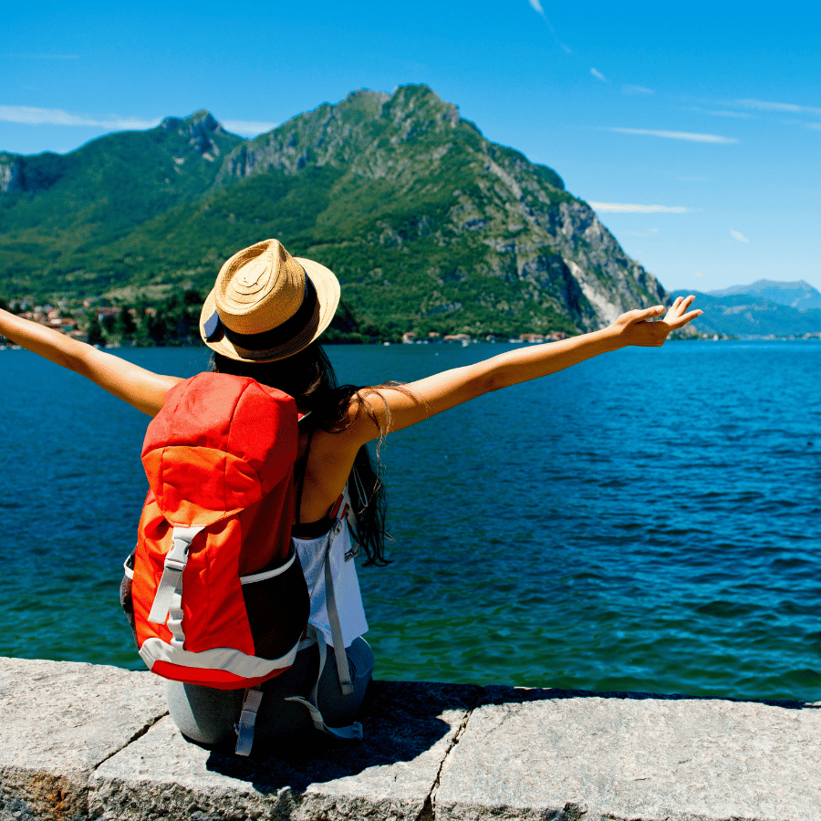 Traveler with red backpack and straw hat, arms outstretched by a blue lake with green mountains.
