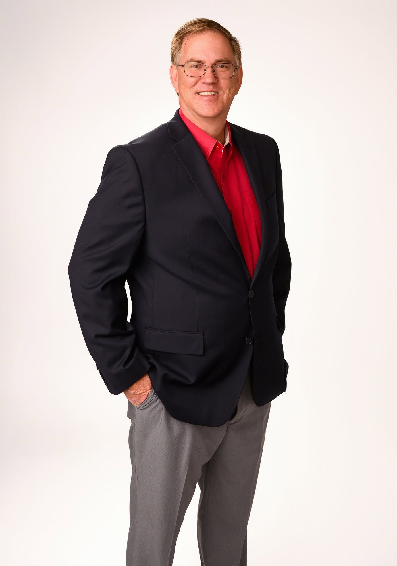 Man in a dark blazer and red shirt, standing against a white studio backdrop, hands in pockets.