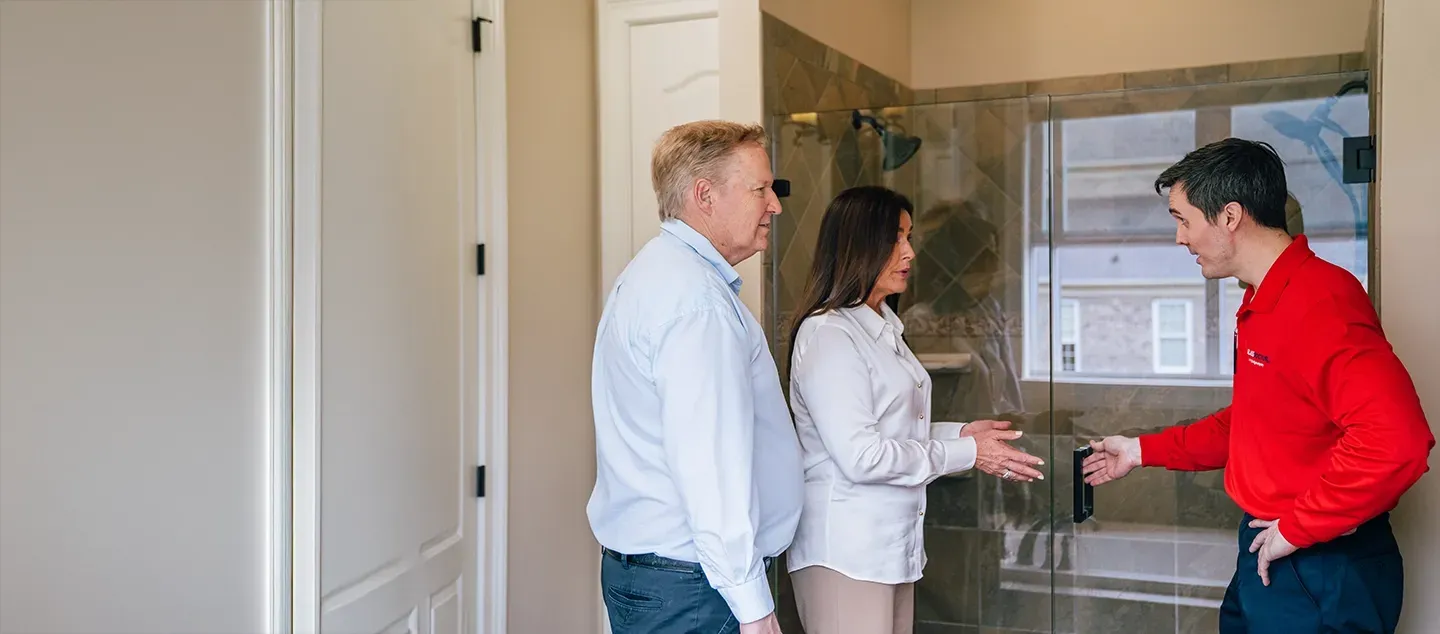A bathroom with three people: two observing a shower with a service person in a red shirt.