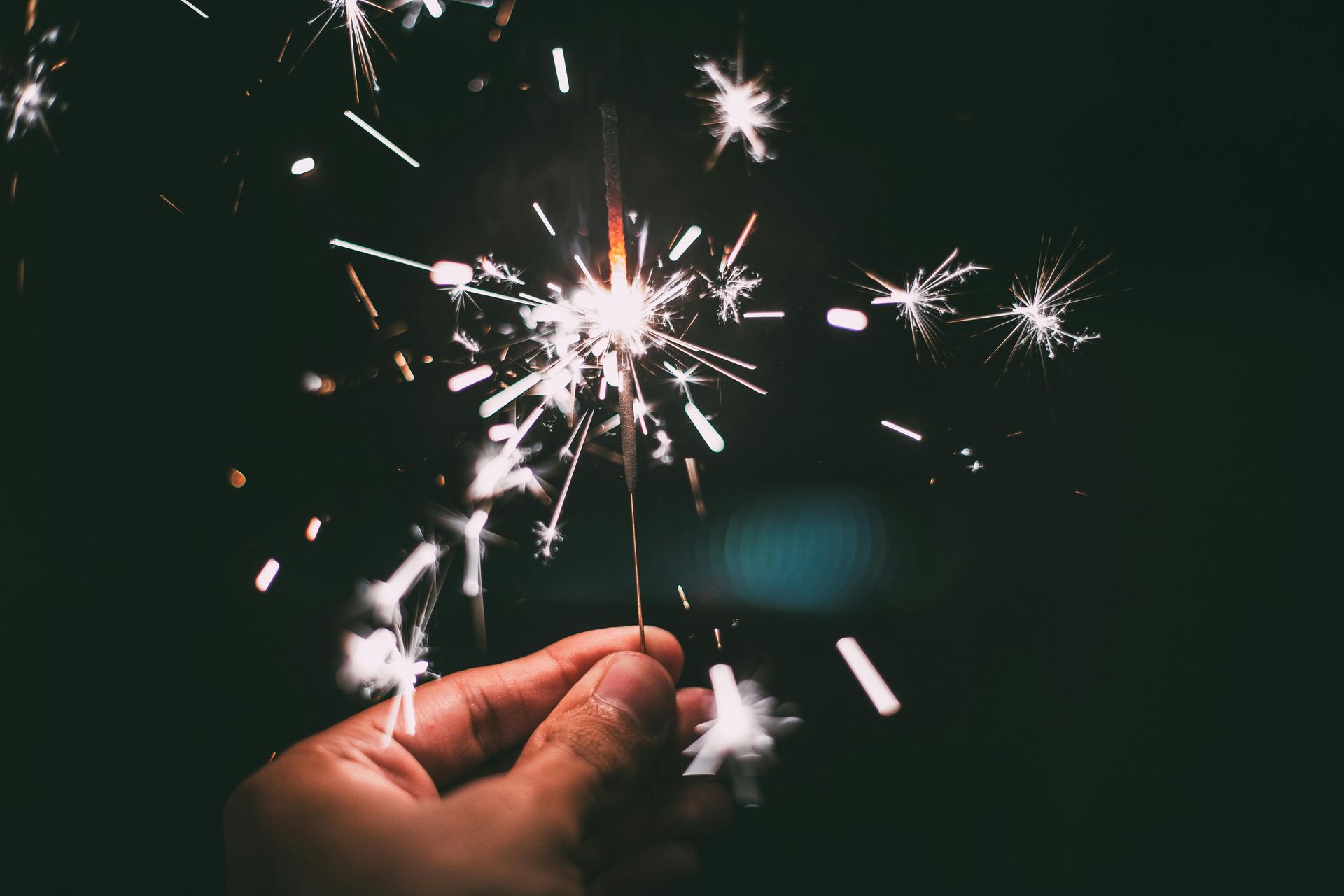 Hand holding a lit sparkler, sparks radiating outwards against a dark background.