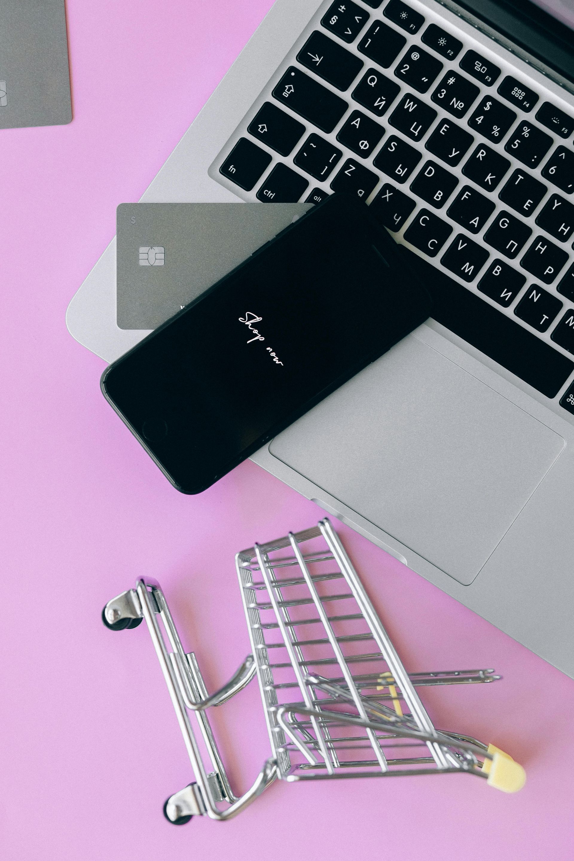 Laptop on pink desk with a credit card, smartphone, and small shopping cart.