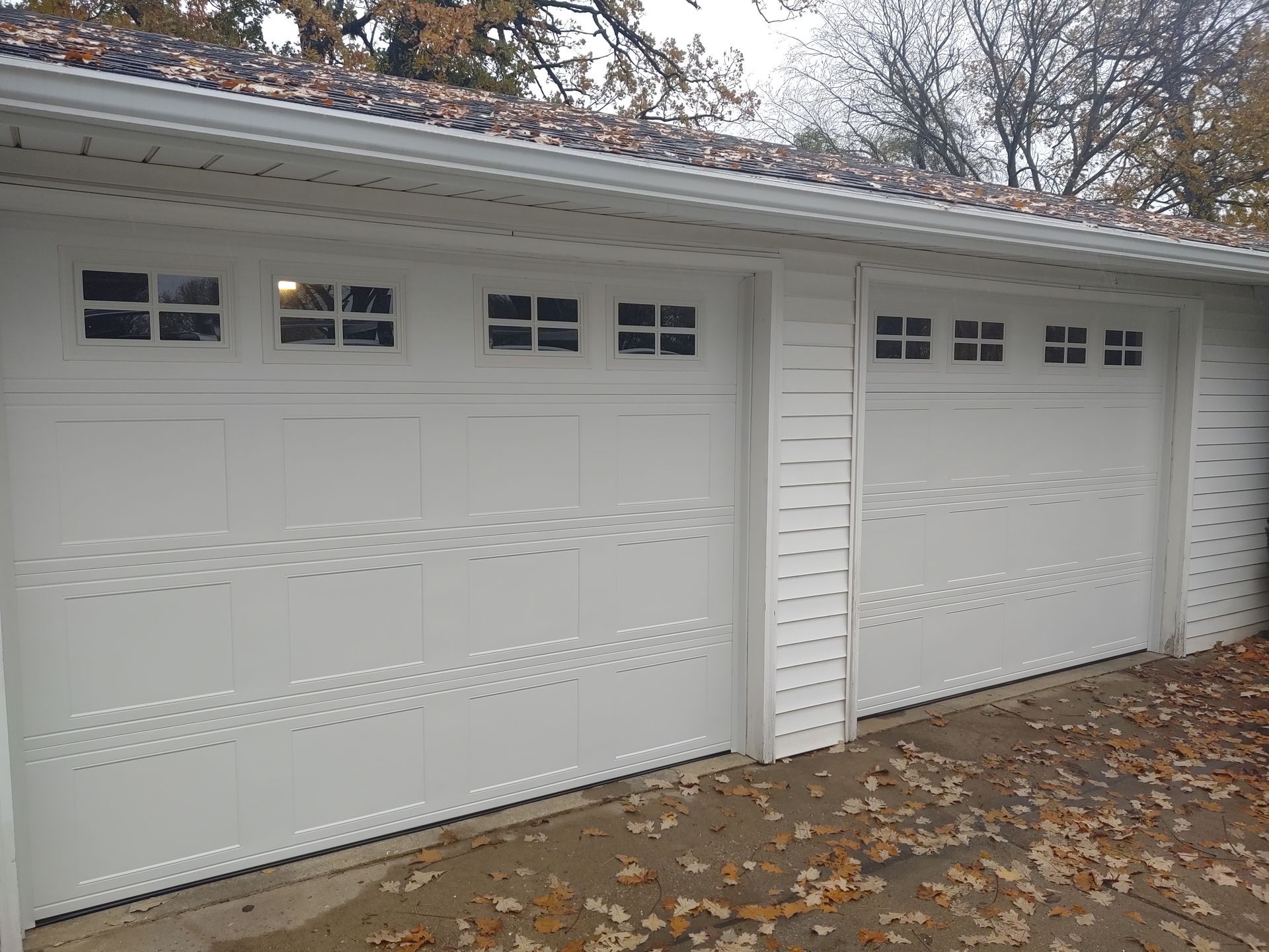 Two white garage doors on a house, with autumn leaves on the driveway.