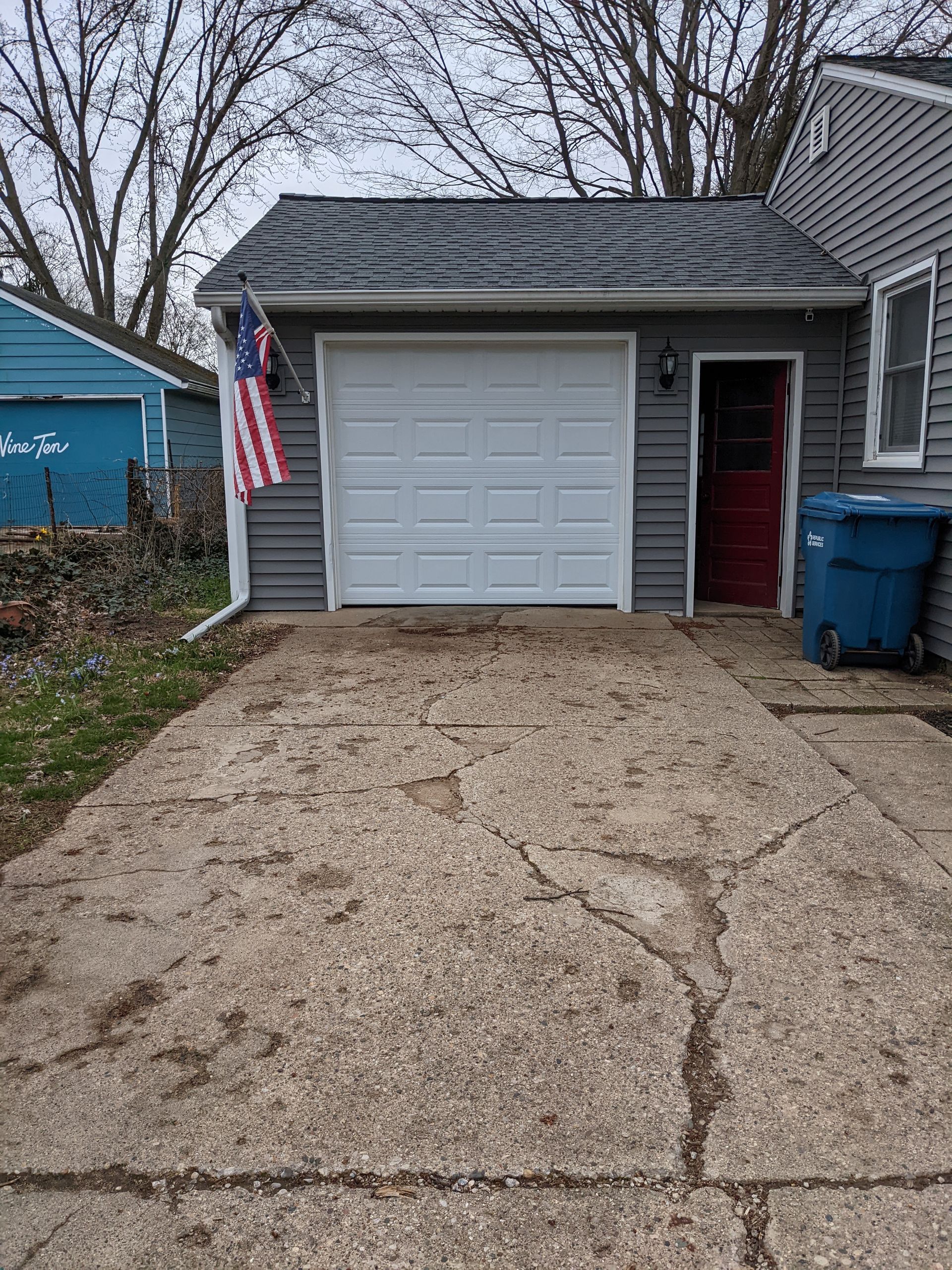 Small house with a single-car garage, gravel driveway, American flag, and blue trash bins.