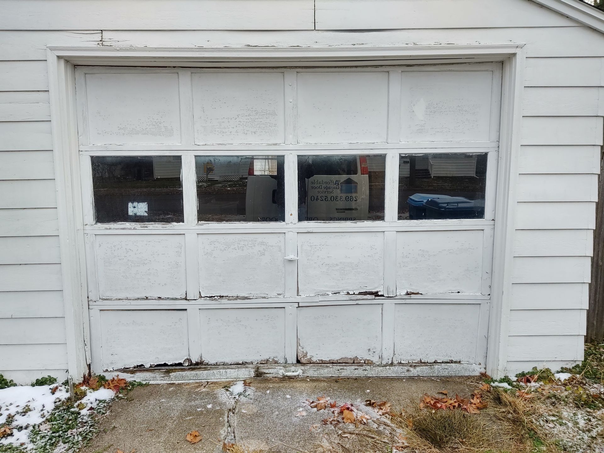 White garage door with four small windows, set in a white-sided exterior wall with a snowy driveway.