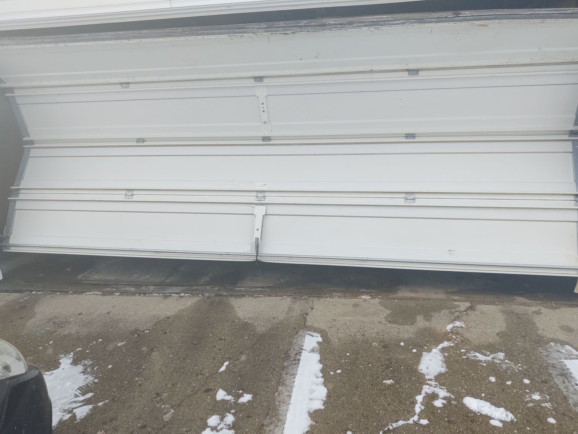 Snow-dusted white garage door with a small dent and muddy ground in front