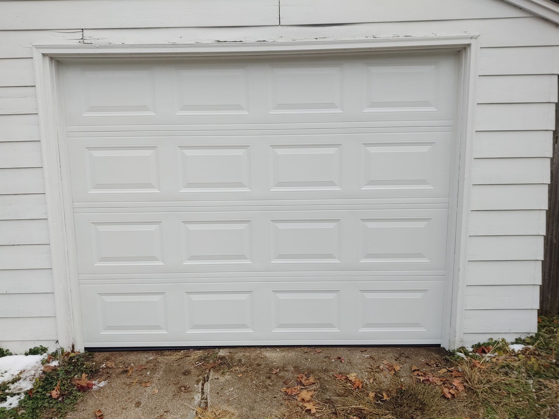 White garage door with rectangular panels on a house exterior, surrounded by white siding and fallen leaves.