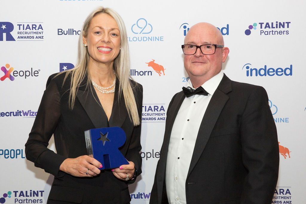 Woman and man holding an award with logos in background. Woman in black blazer, man in tuxedo.