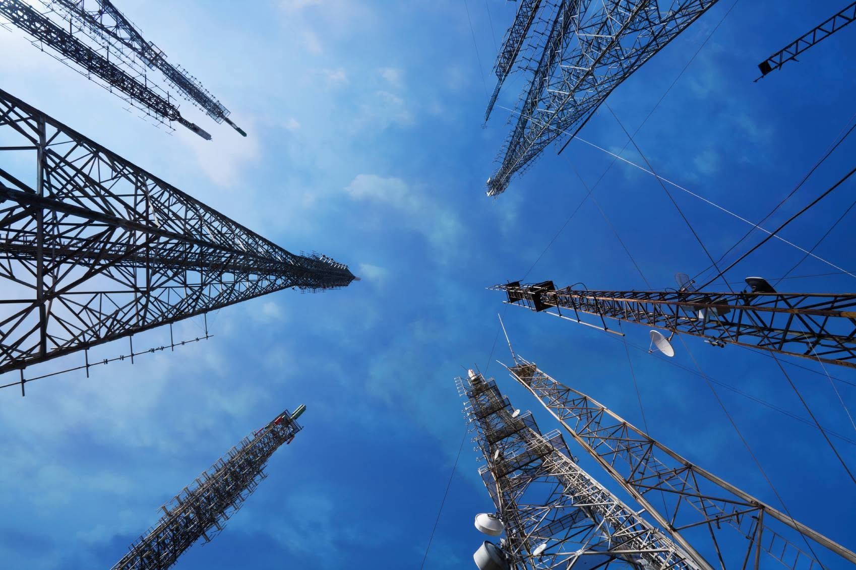 Metal communication towers viewed from below against a bright blue sky with clouds