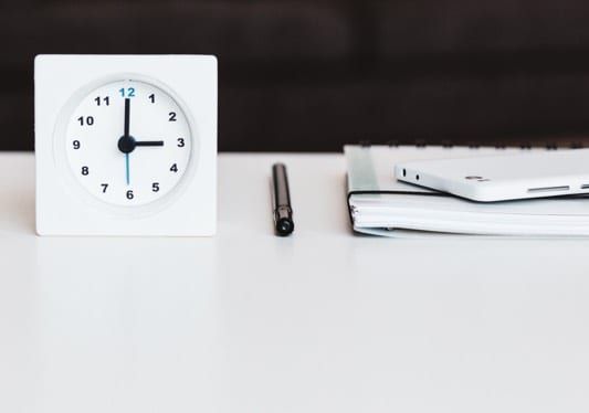 White square clock at 3 o'clock, pen, notebook, and phone on a white table.