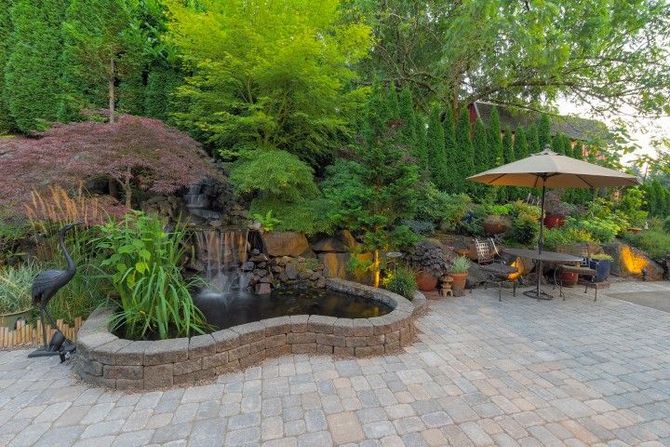 A backyard patio with a stone-walled pond, a small waterfall, lush landscaping, and a dining table under an umbrella.