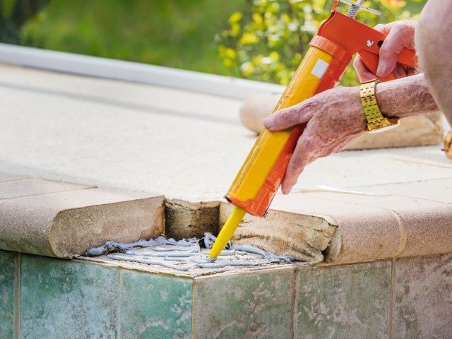 A hand uses a yellow caulk gun to apply sealant to the gap between concrete pool coping tiles.