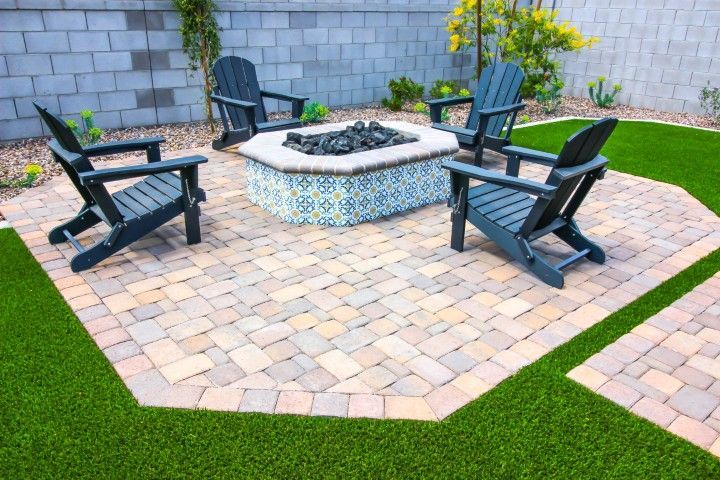 Four dark patio chairs surround an octagonal tiled fire pit on a paver patio set in a backyard with artificial turf.