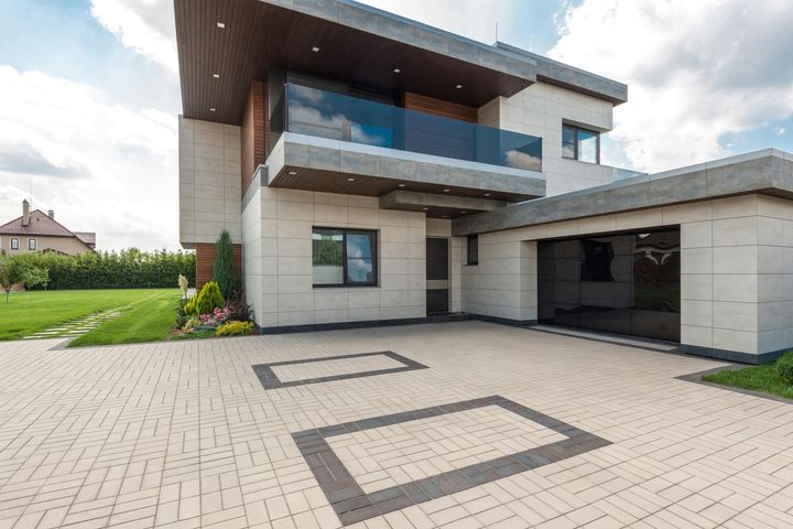 A modern, two-story house with beige stone siding, a dark roof, a balcony, and a paved driveway with dark stone accents.