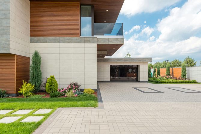 Modern house exterior with wood accents, cream stone panels, a glass balcony, and a paved driveway under a blue sky.
