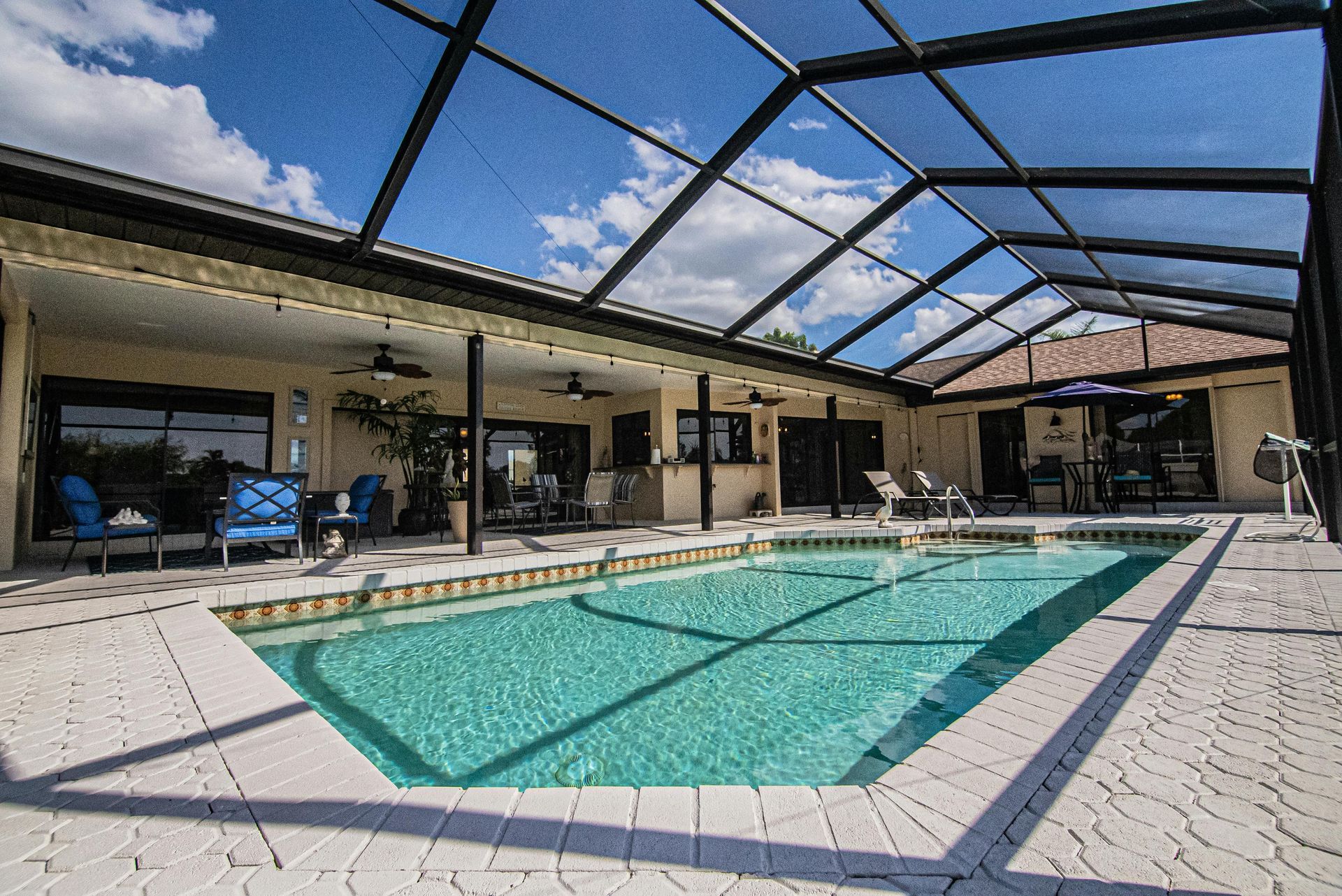 A bright swimming pool inside a screened-in lanai attached to a house under a blue sky with scattered clouds.