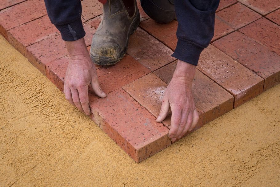Hands place red rectangular paving bricks onto a flat, yellow sand surface.