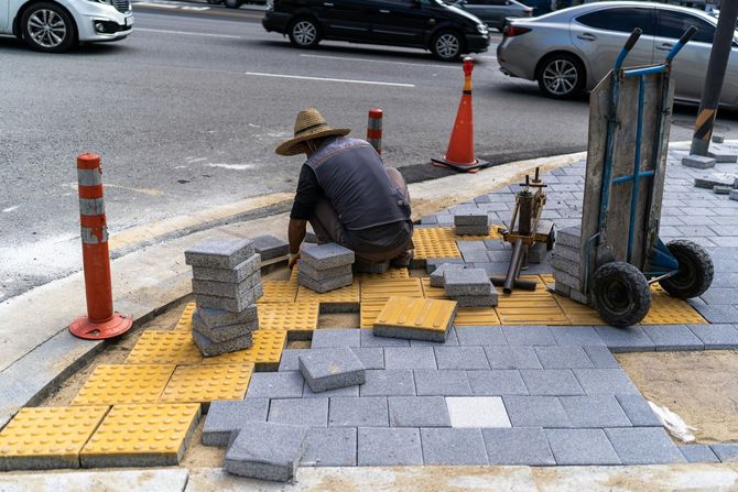 A worker in a straw hat installs yellow tactile paving and gray stone pavers on a sidewalk next to a road.