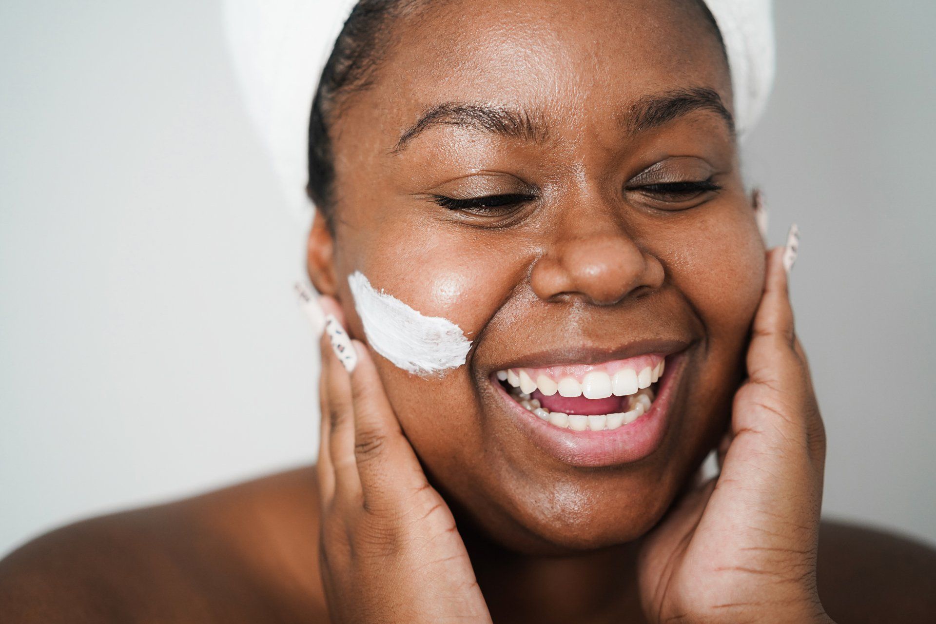 A woman with a towel wrapped around her head is smiling while applying cream to her face.