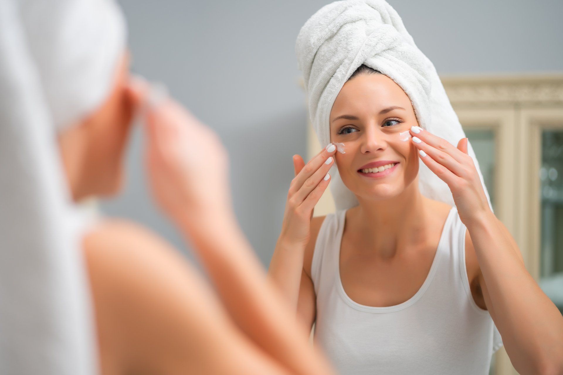 A woman with a towel wrapped around her head is looking at her face in the mirror.