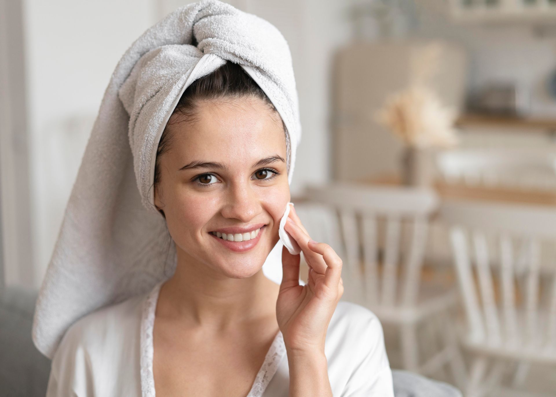A woman with a towel wrapped around her head is cleaning her face with a cotton pad.