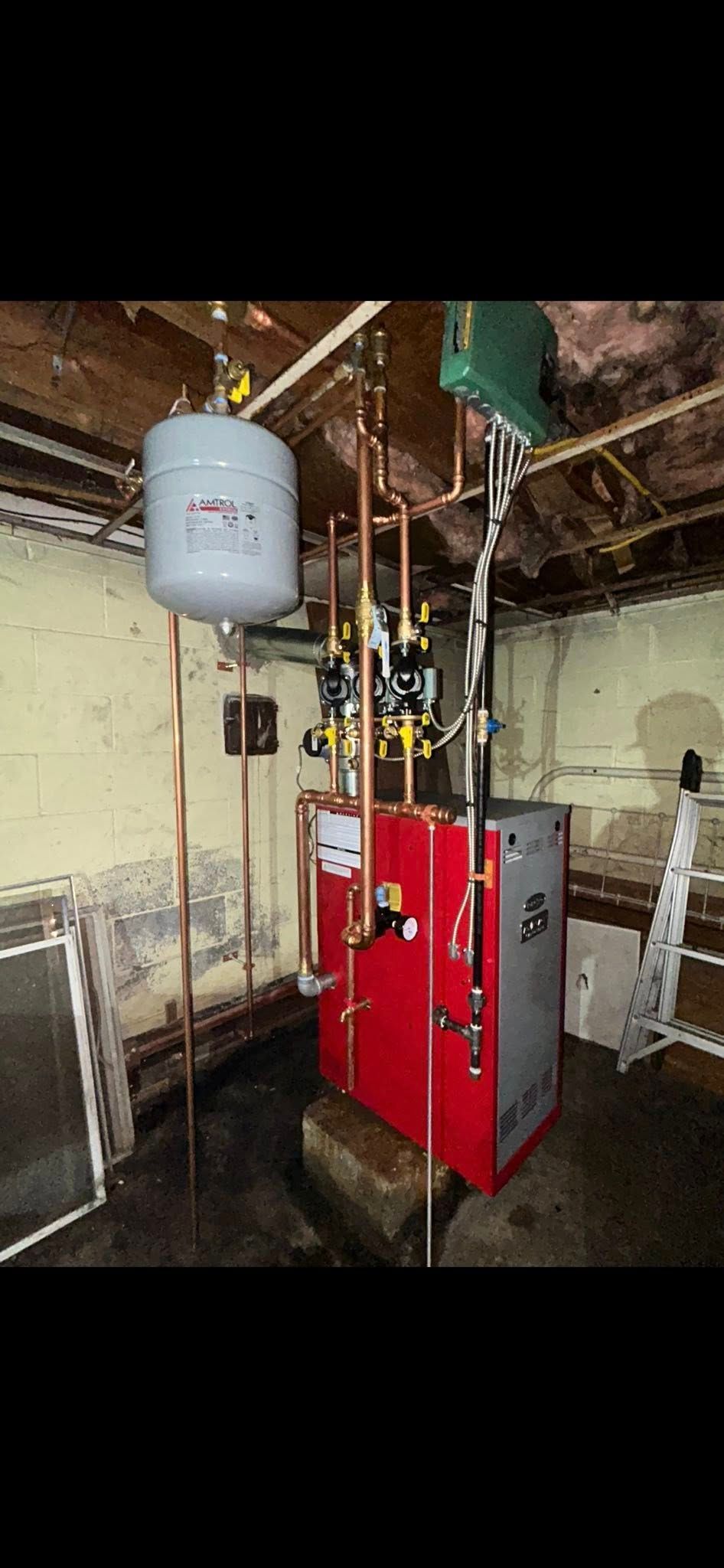 A red boiler and grey expansion tank connected by copper pipes in an unfinished basement with concrete walls.