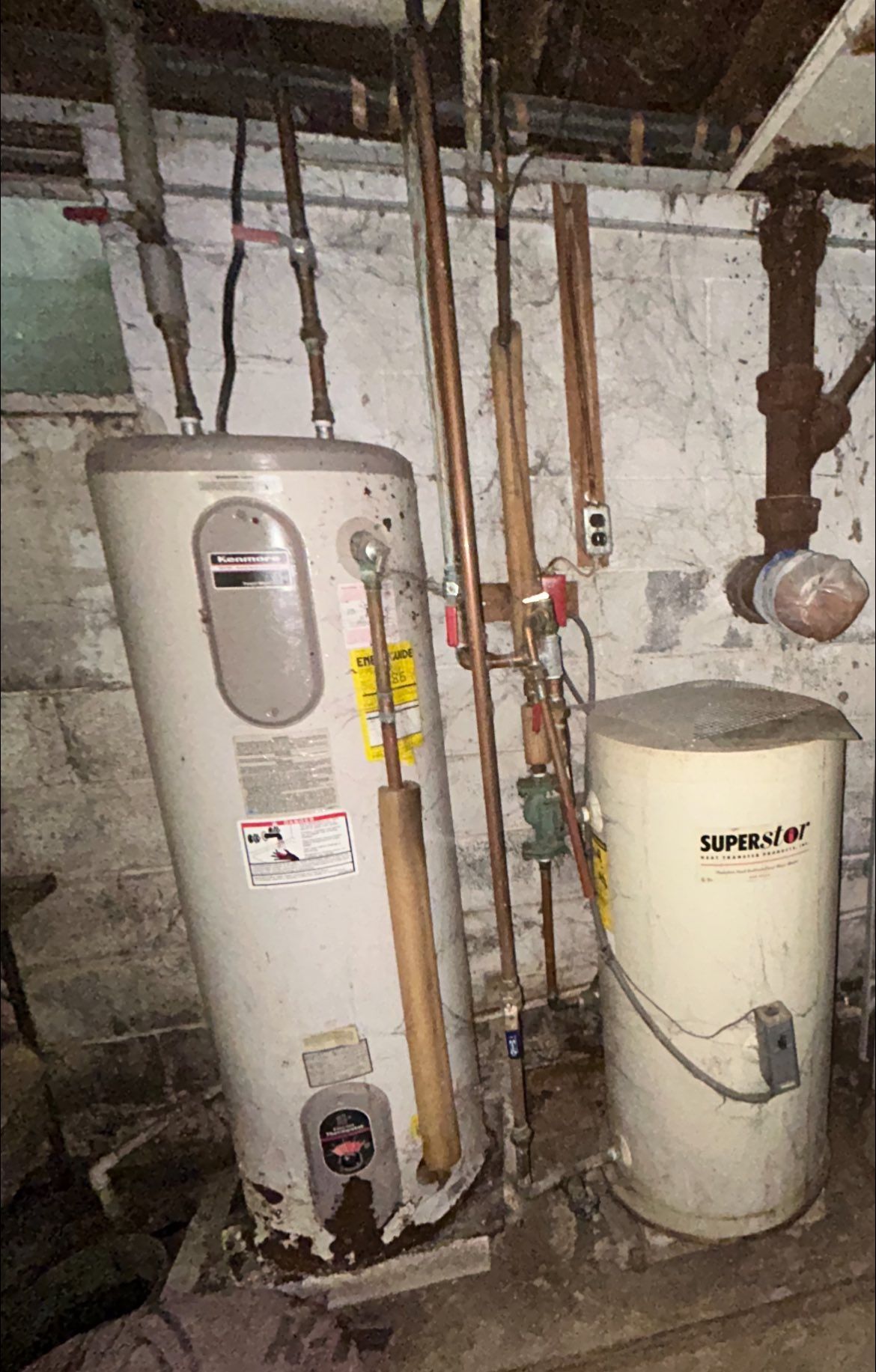 A water heater and a secondary storage tank sit on a basement floor with exposed pipes against a concrete wall.