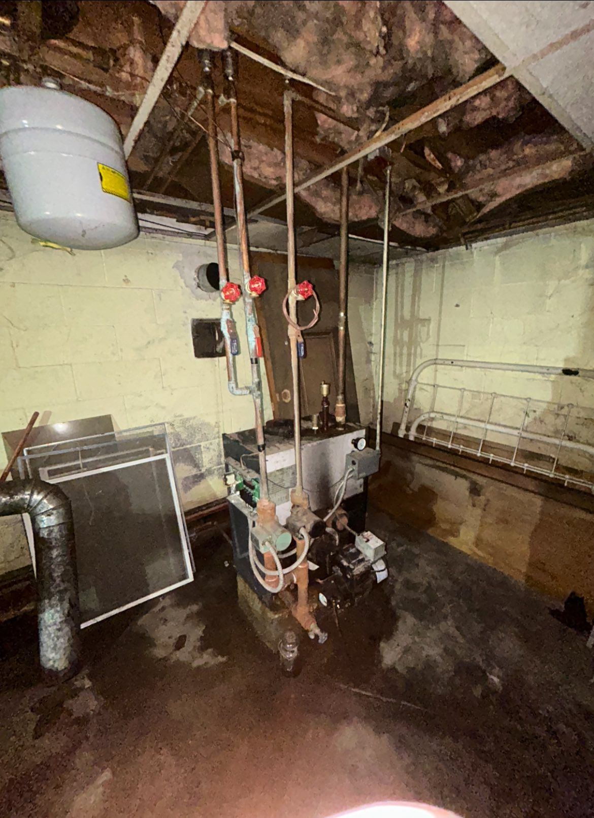 A residential boiler in a basement with exposed copper piping, an expansion tank, and a damp floor.