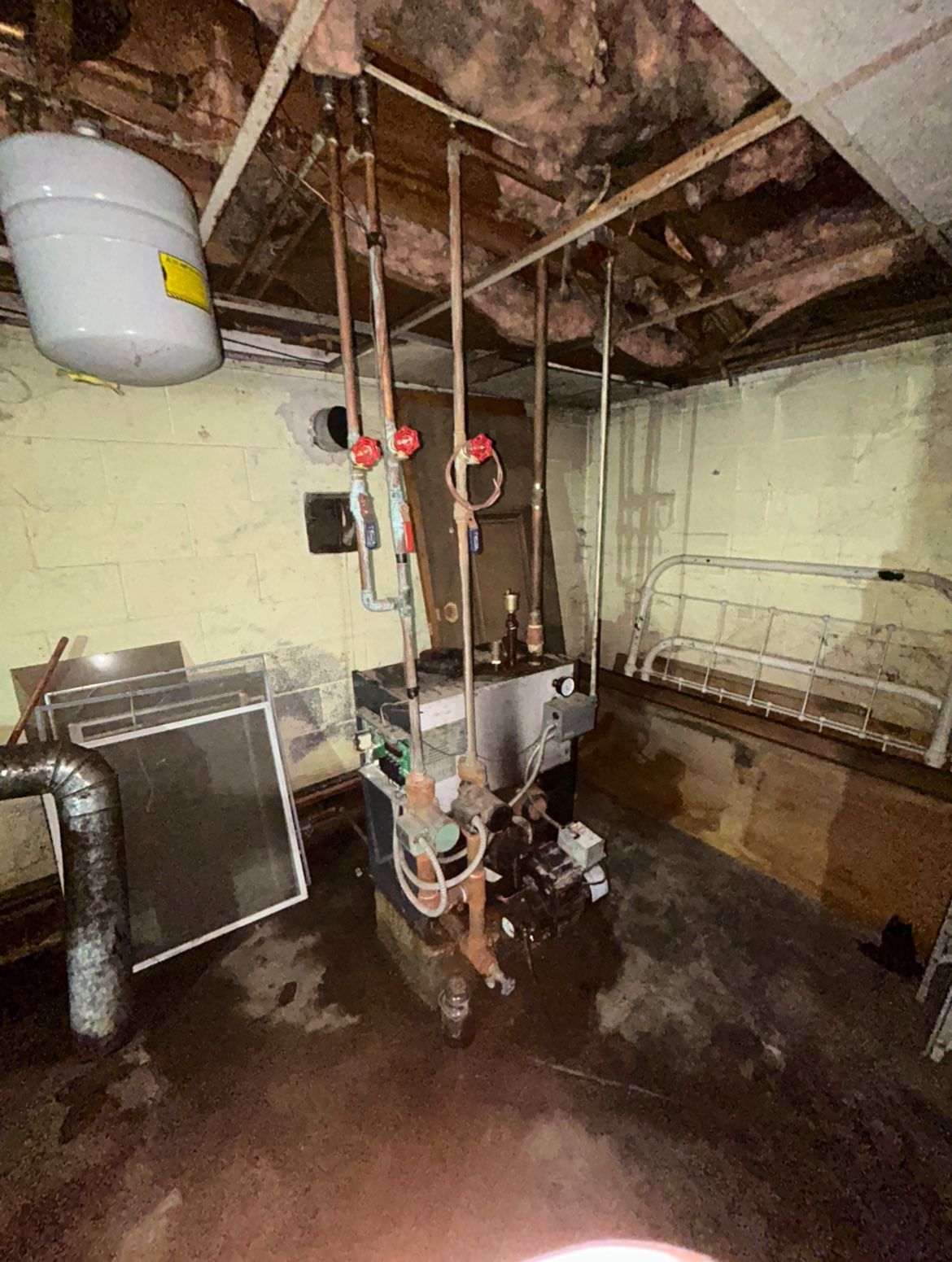A boiler sits in a basement with exposed insulation overhead, copper piping, and a white expansion tank on the wall.