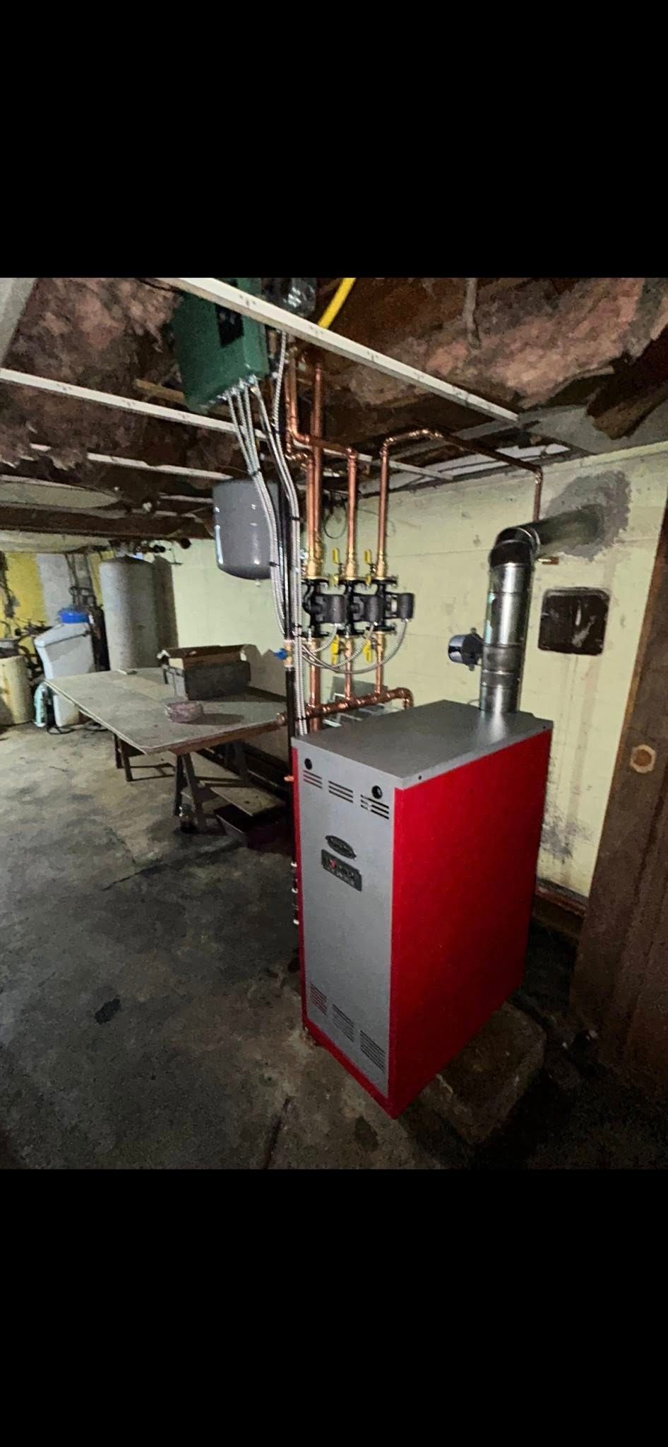 A red and silver boiler with attached copper piping stands in an unfinished basement with exposed ceiling joists.