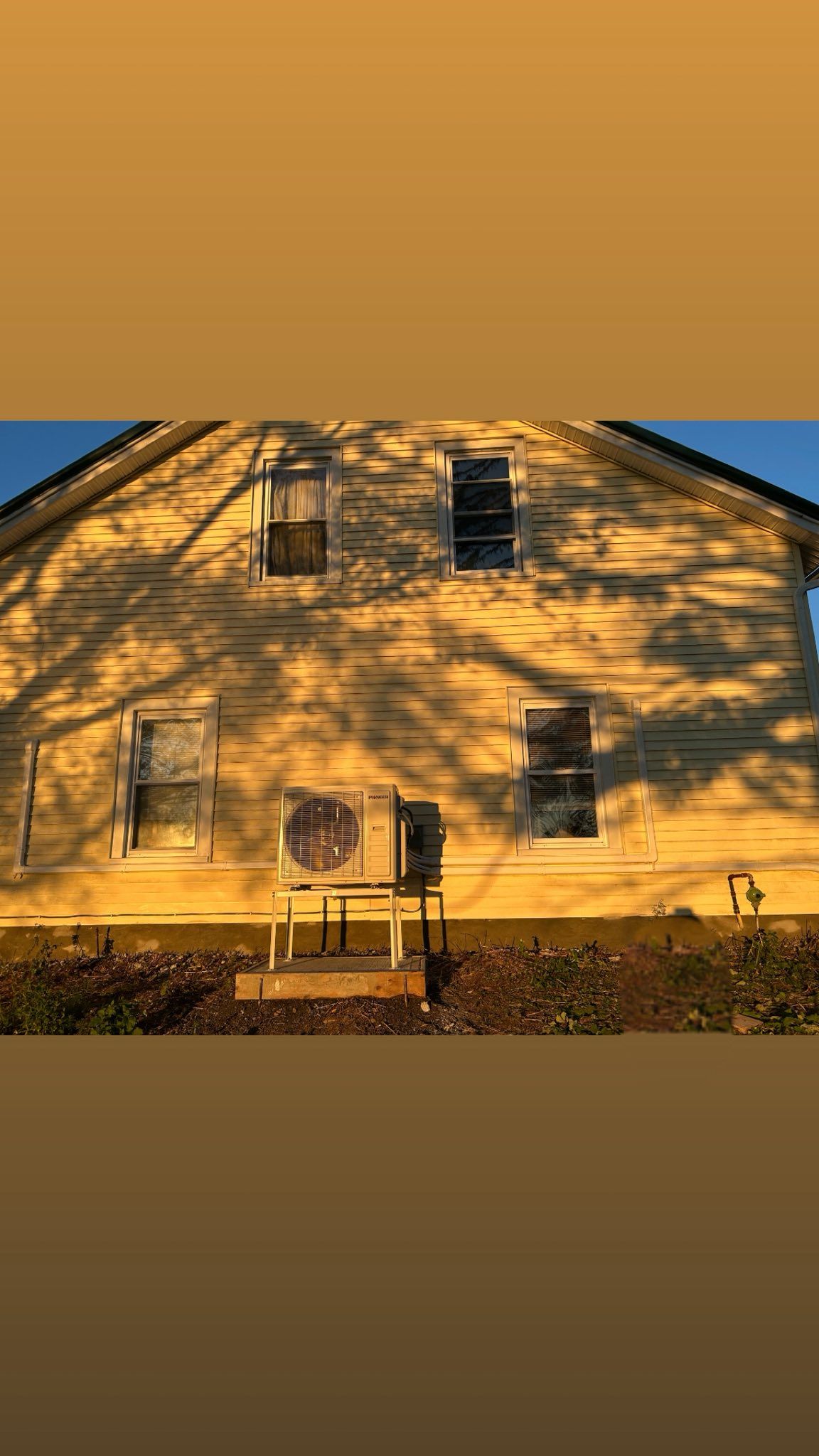 A pale yellow house exterior at sunset, with four windows and an outdoor air conditioning unit in front.