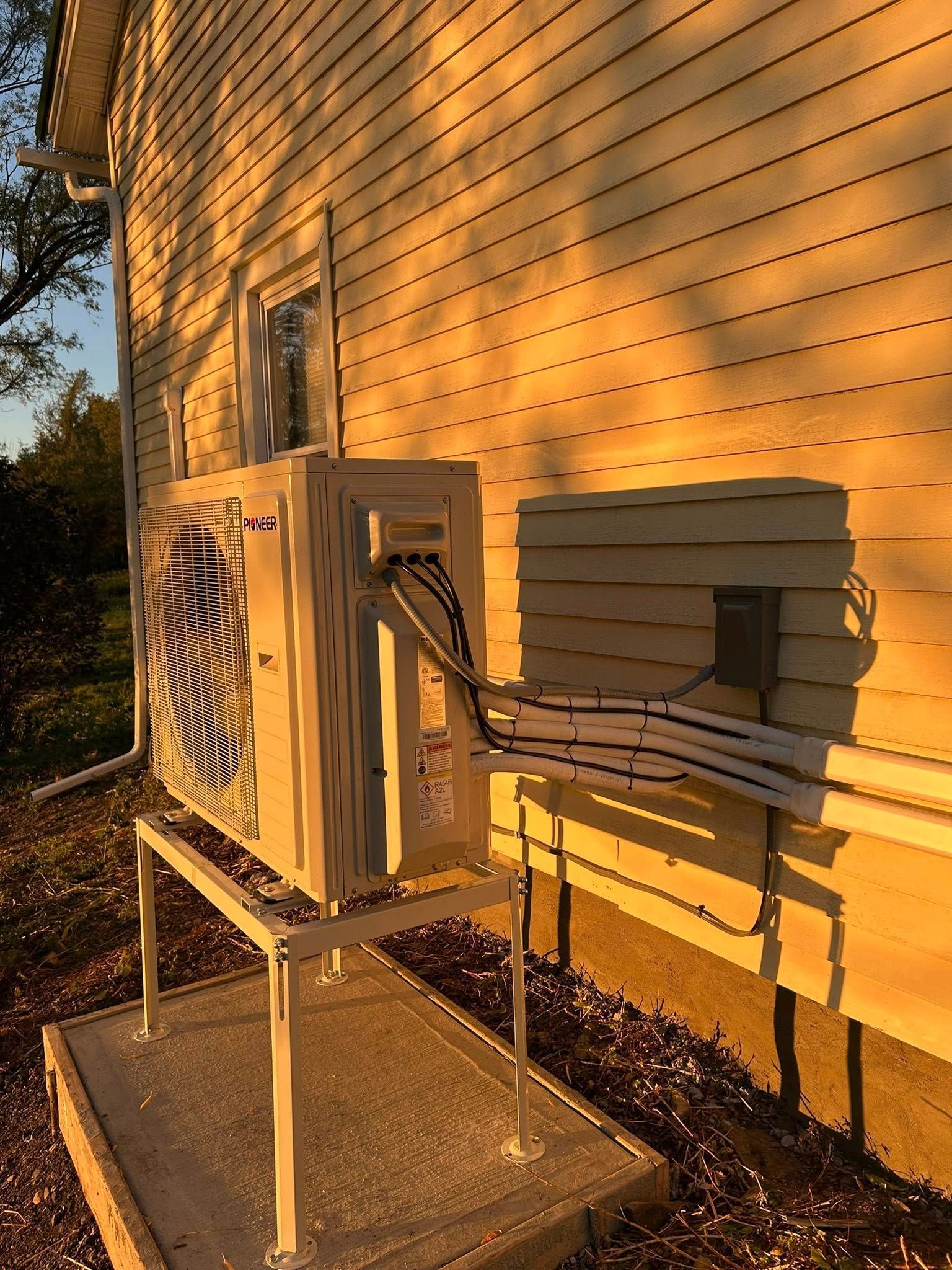 An outdoor heat pump unit sits on a raised metal stand on a concrete pad against a yellow, vinyl-sided house at sunset.