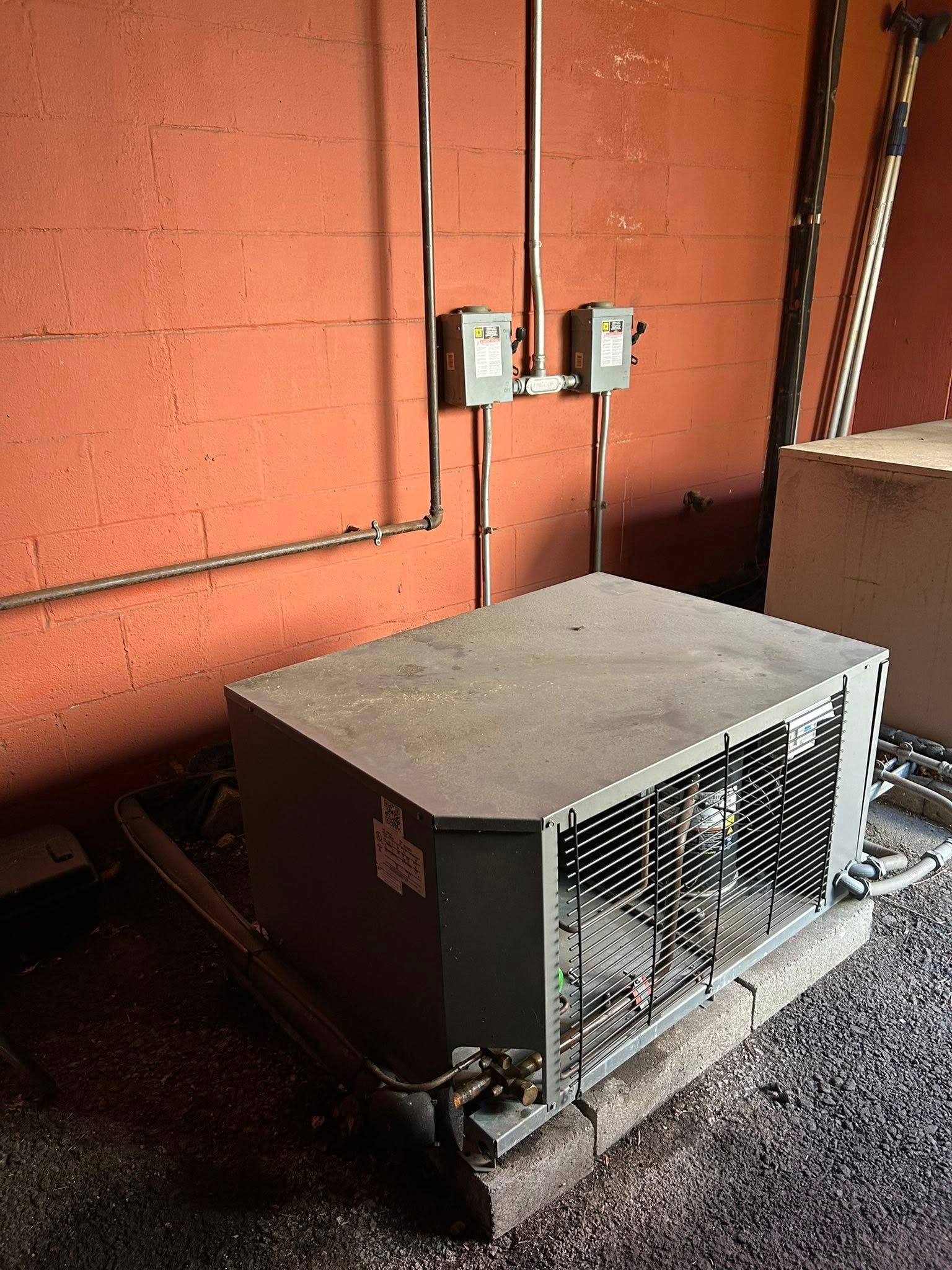 An industrial outdoor HVAC unit sits on a concrete pad against a textured orange brick wall with two electrical boxes.