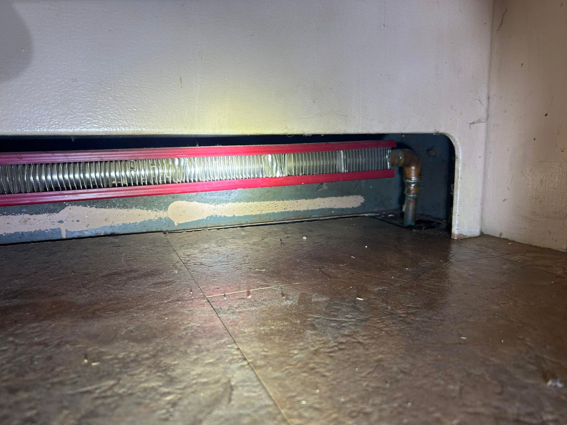 A metal heating element with a red-taped edge is installed along the base of a wall above a wood-look floor.