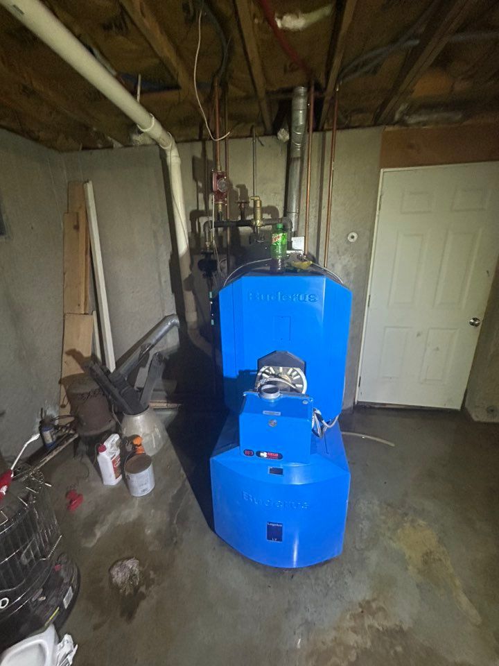 A blue oil-fired residential boiler stands in a basement with concrete walls, exposed ceiling joists, and pipes above.