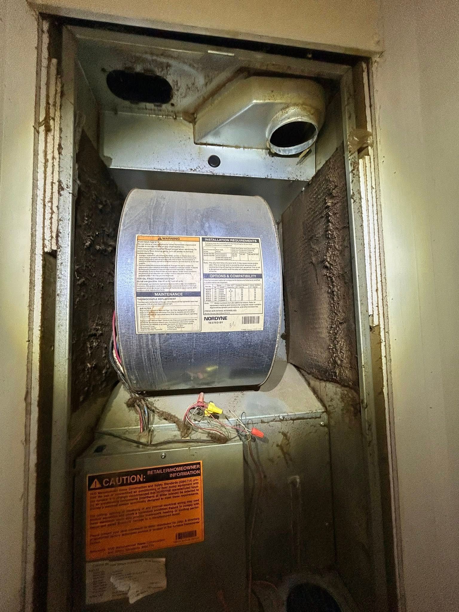 An open utility closet housing a dusty HVAC furnace unit with visible internal components, wiring, and warning labels.