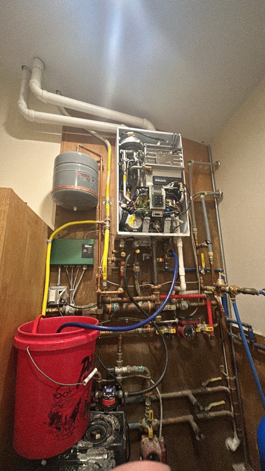 A wall-mounted tankless water heater with exposed pipes and a red bucket beneath, indicating a maintenance flushing process.