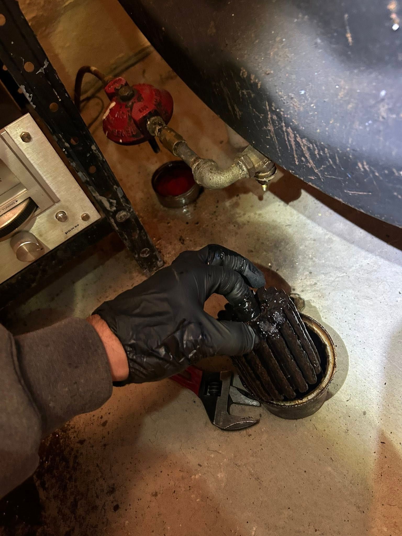 A gloved hand holds a dirty oil furnace filter over a drain, with a nearby red pump visible on the concrete floor.