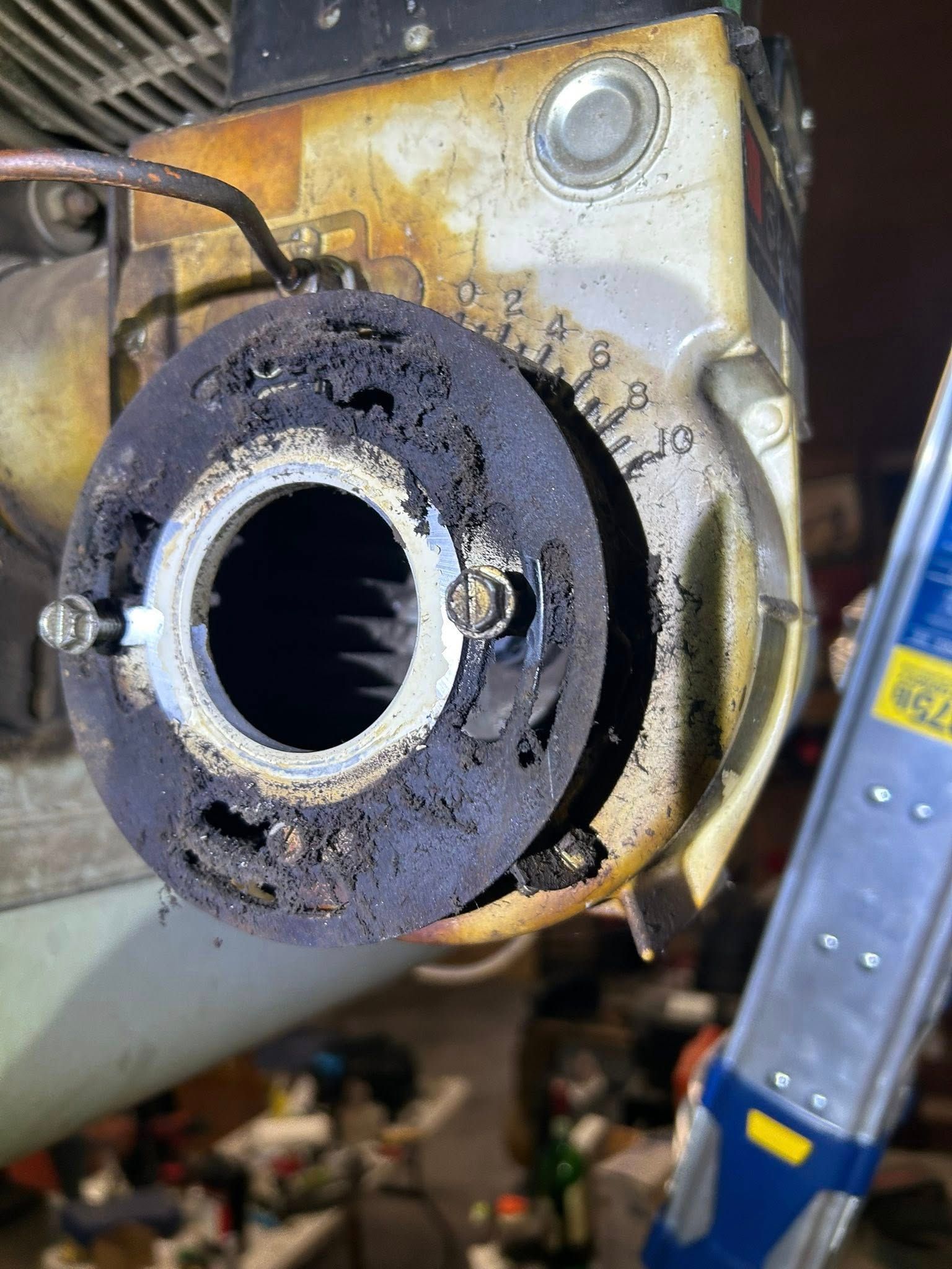 A dirty, circular mechanical component of an oil furnace burner covered in soot, with a ladder visible to the right.