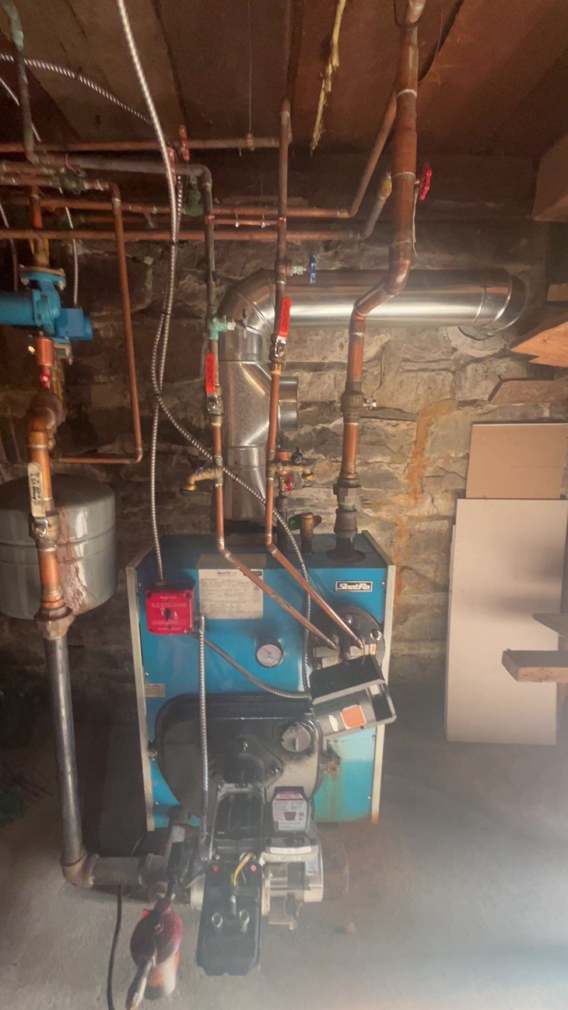 Blue oil-fired residential boiler with copper plumbing and silver flue pipe in a stone-walled basement.
