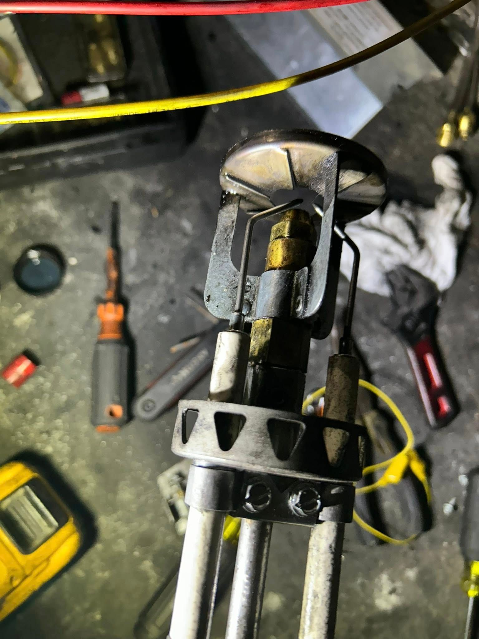 A close-up view of a metal furnace ignition assembly with wires and a circular burner top, held in a cluttered workspace.
