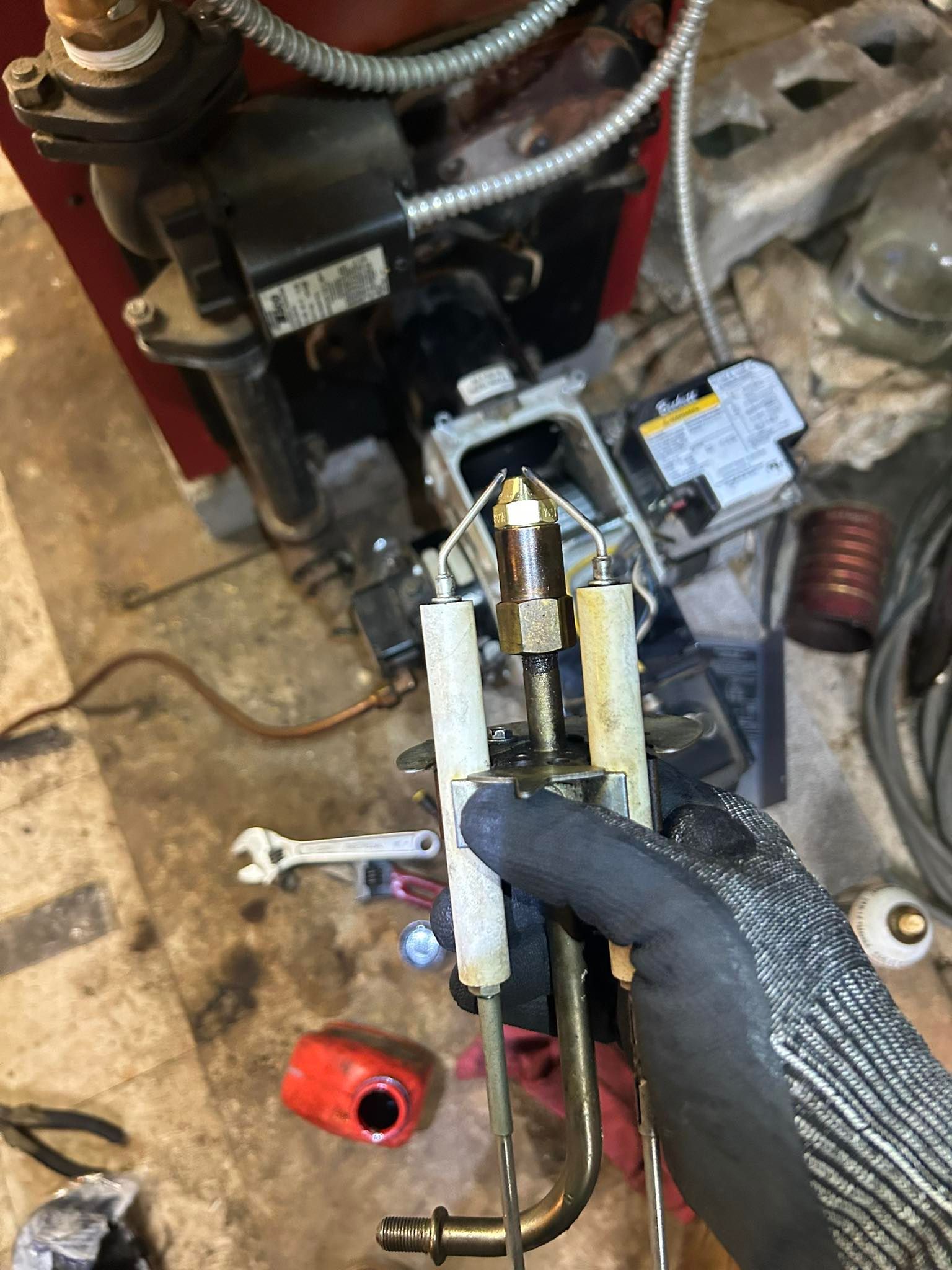 A gloved hand holds a residential oil burner nozzle assembly with ceramic ignition electrodes in front of a furnace.