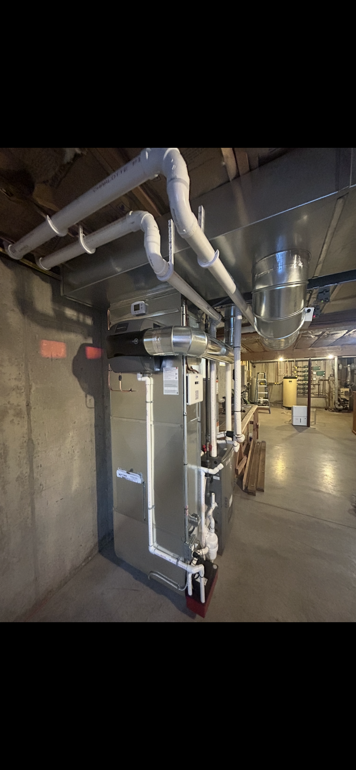 A tall, metallic HVAC furnace unit with white PVC piping installed in a basement with concrete walls and exposed joists.