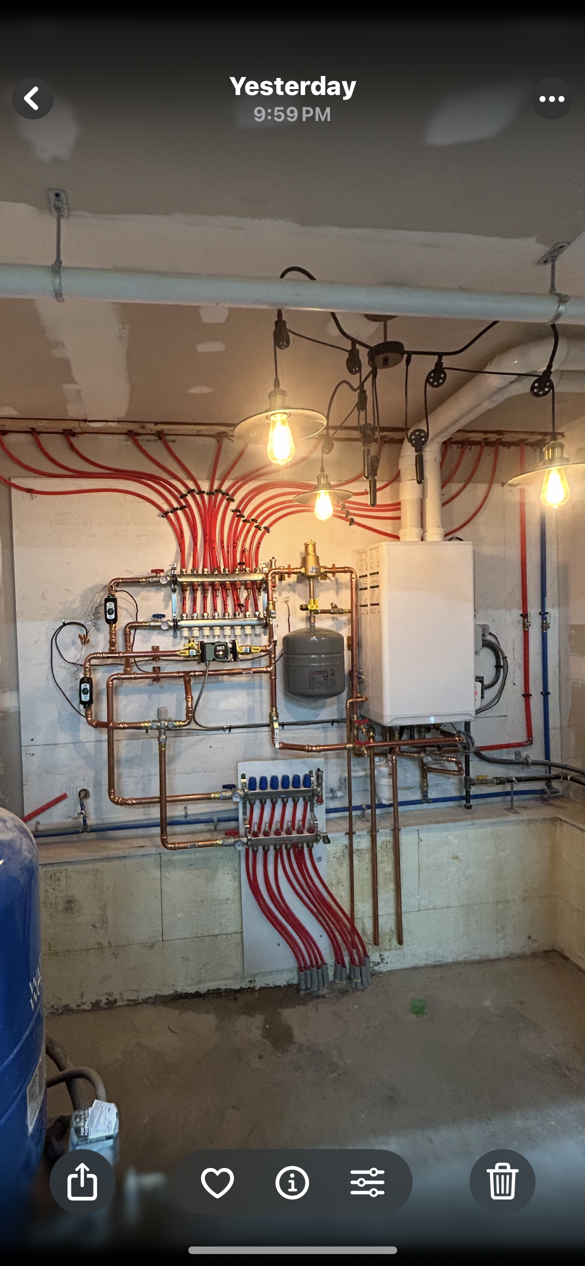 A wall-mounted boiler and manifold system featuring numerous red and blue pipes installed against an unfinished basement wall.