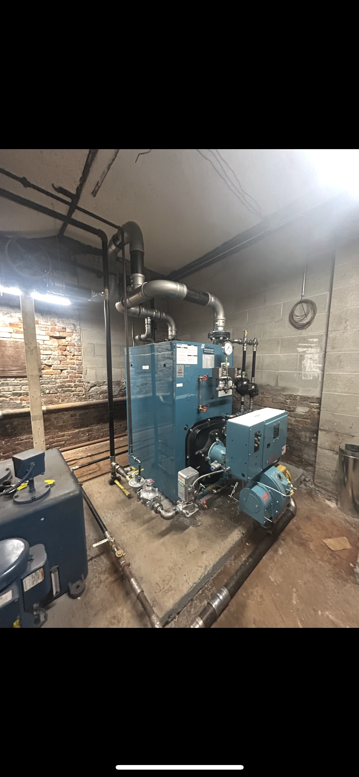 A blue industrial boiler with pipes sits on a metal platform inside a concrete basement utility room.